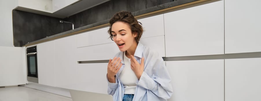 Portrait of young freelance girl connects to online meeting, video chats on laptop, sits on kitchen floor and enjoys her conversation.