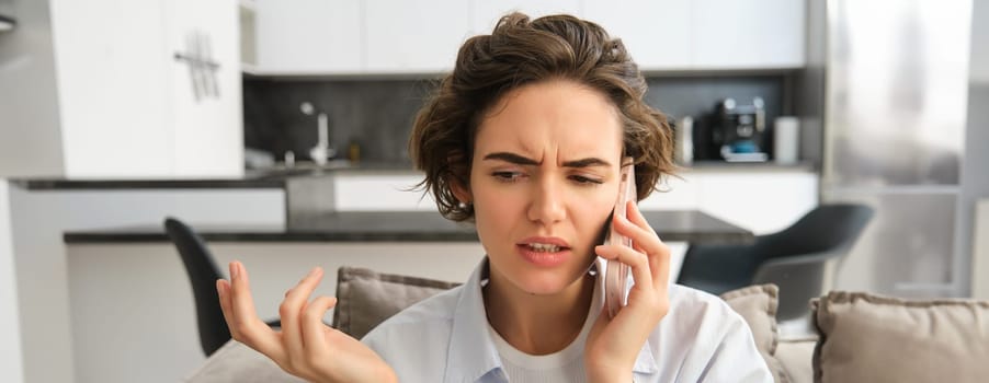 Close up portrait of confused young woman answers phone call and shrugs, looks puzzled while listens to caller, having complicated conversation over the telephone.