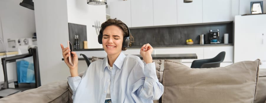 Portrait of beautiful, happy young woman, listening music in headphones, holding smartphone and dancing on sofa at home, enjoys spending time indoors alone.