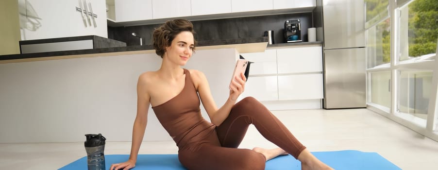 Young fitness instructor looking at her smartphone, sitting on rubber mat at home. Young woman doing workout training, looking at exercise videos on mobile app.