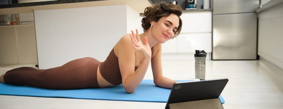 Sport and lifestyle. Young fitness woman, gym instructor waves hand at tablet, joins online video class, yoga and mindfulness workout, training from home indoors.