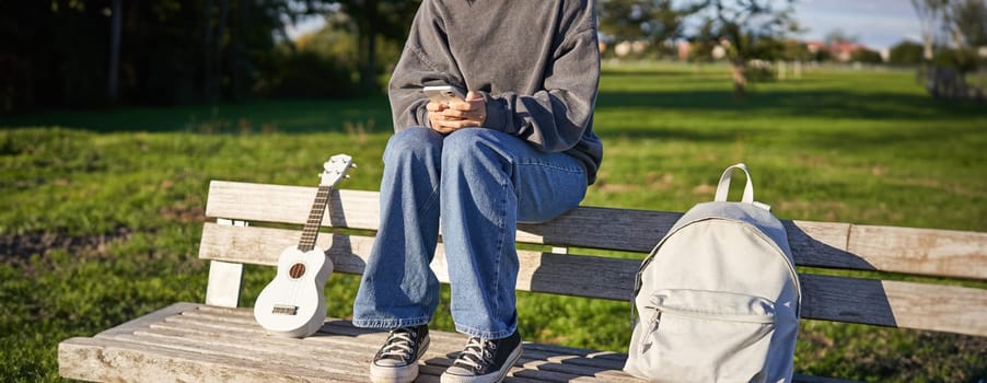 Cropped shot of teen girl body, sitting on bench with ukulele, using smartphone, hands holding mobile phone.