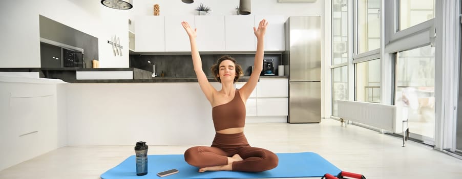 Smiling young woman in activewear, sitting on mat at home, raising arms up while meditating, practice yoga, doing mindful workout. Sport and wellbeing