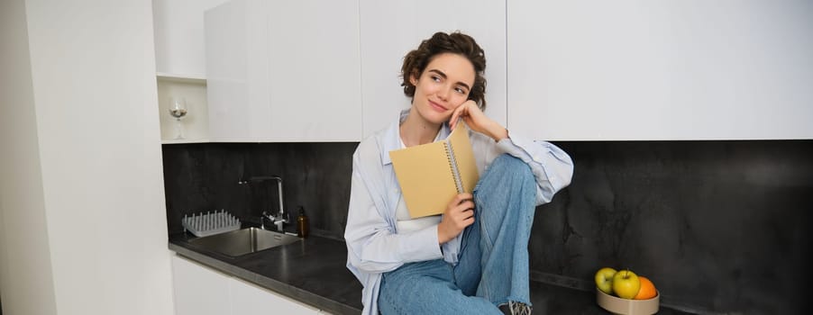 Lifestyle and people concept. Young woman enjoying weekend at home, reading notes in her journal, sits on kitchen counter with notebook and smiling.