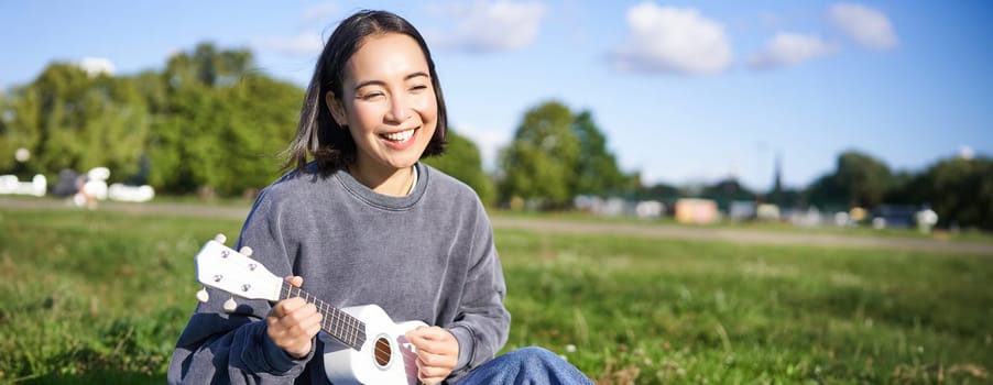 Carefree asian girl singing and playing ukulele in park, sitting on grass, musician relaxing on her free time outdoors.