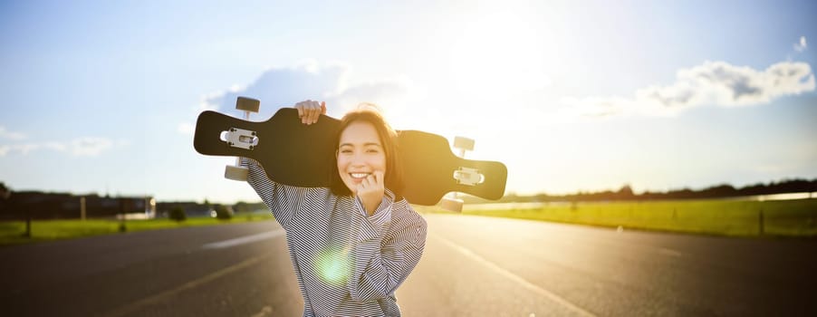 Happy skater girl walking towards the camera, sunbeams shining at lense. Young woman skating on cruiser, holding longboard on shoulders.