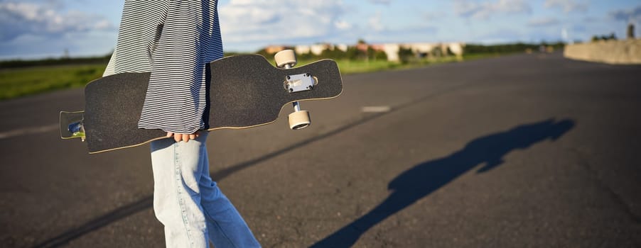 Cropped shot of teen skater girl, hands holding longboard, walking with skateboard on concrete road.