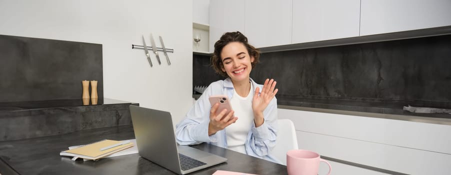 Portrait of young woman video chats with friend on smartphone, waves hand at mobile phone camera, calls someone using application.