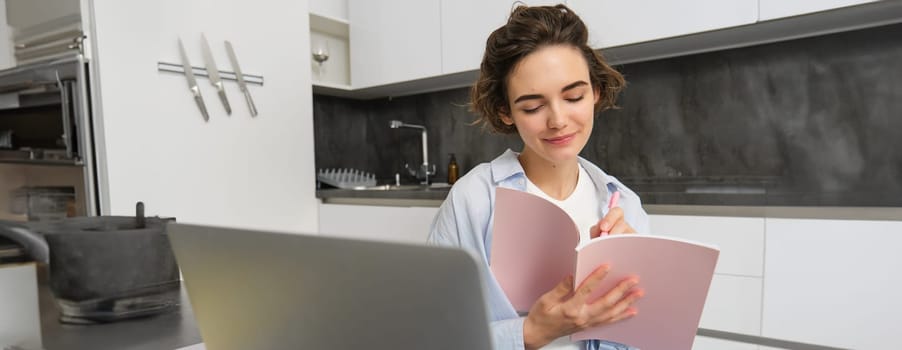 Young woman checking her schedule while working on remote from home, using laptop, looking at her daily planner, making notes, writing down information in notebook.