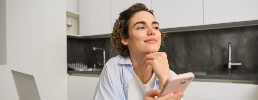 Portrait of modern brunette woman holding mobile phone and smiling. Girl sits in kitchen at home with smartphone, using app to order delivery, does online shopping.