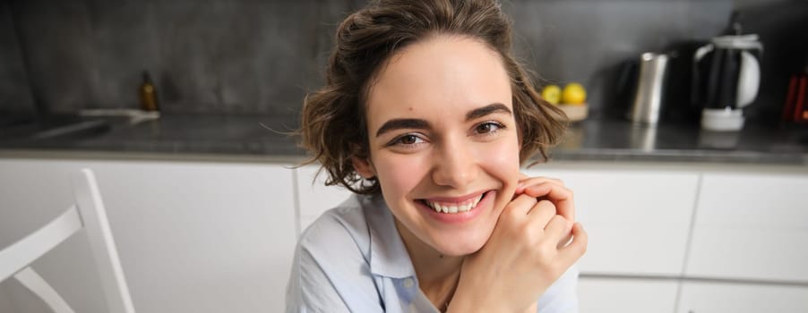 Close up portrait of young woman, 25 years old, sitting in her kitchen alone, express candid happiness, smiles, leans head on hands.