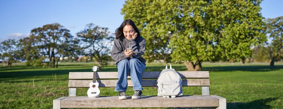 Young woman with ukulele, sitting on bench in park, using mobile phone app, holding smartphone and smiling.