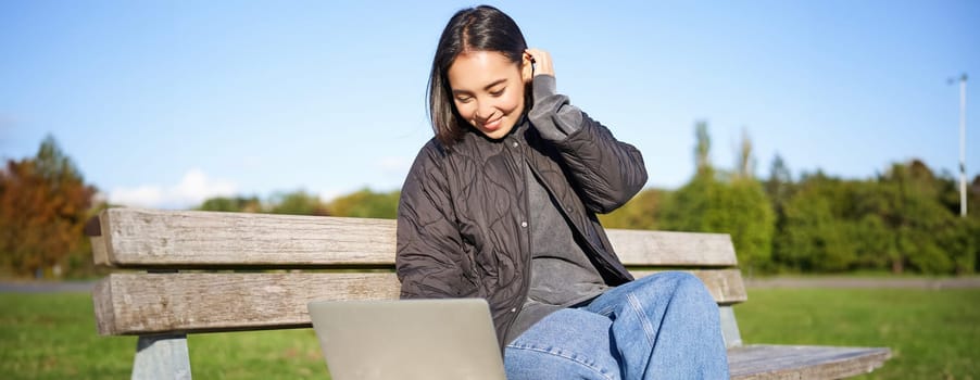 Young brunette girl sits in park with laptop, watches video or browses internet, rests outdoors on bench in sunny park.