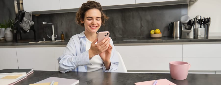 Technology and lifestyle. Young woman sits at home, uses smartphone in her kitchen and smiles.
