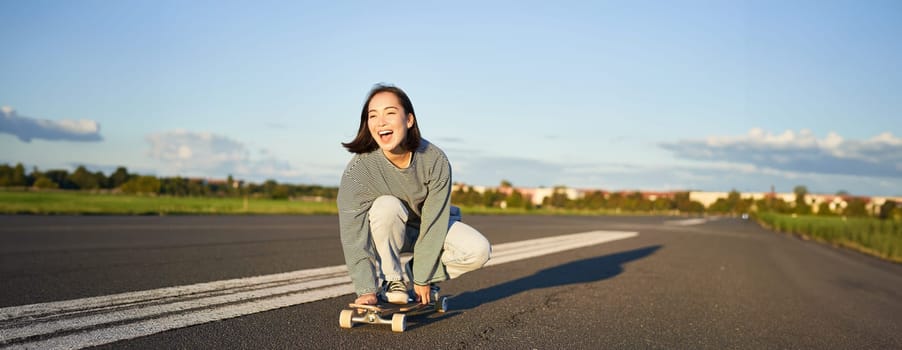 Freedom and happiness. Happy asian girl riding her longboard on an empty sunny road, laughing and smiling, skateboarding.