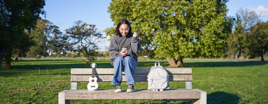 Excited girl looks at smartphone and celebrates, wins on mobile phone, sits with ukulele and backpack in park on bench on sunny day.