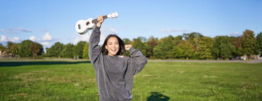 Upbeat young woman dancing with her musical instrument. Girl raises her ukulele up and pose in park on green field.