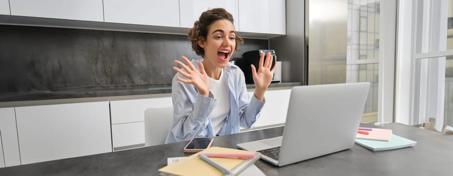 Friendly smiling woman sists at home, waves hands at laptop camera, says hello, joing online meeting, connects to video chat.