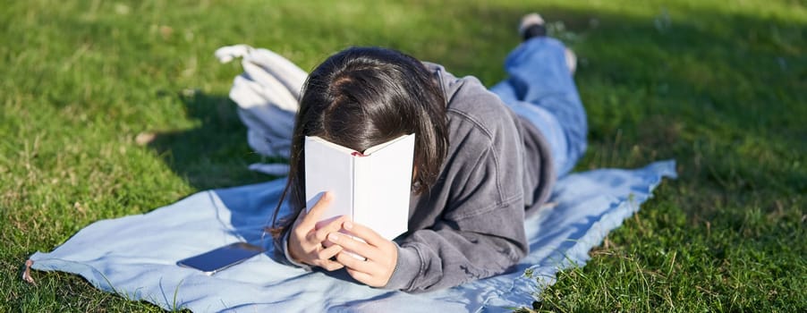 Portrait of happy girl laughing, covering face with her book, lying on grass in park. Copy space