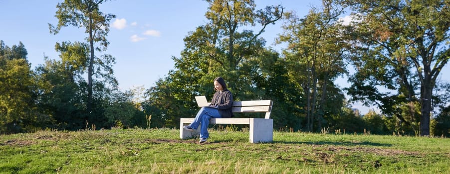 Side view of young woman sitting alone in park on bench, using her laptop to study or work remotely from outdoors.