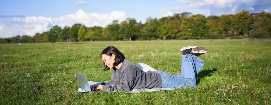 Portrait of young happy asian girl lying on blanket in park, watching videos and browsing internet on her laptop.