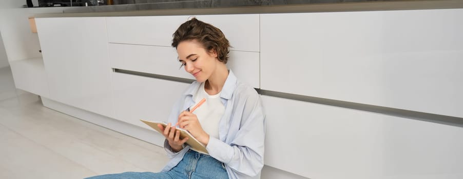 Portrait of young creative girl, student does her homework, writes in diary, holds journal and makes sketches, draws in planner, sits on floor at home.