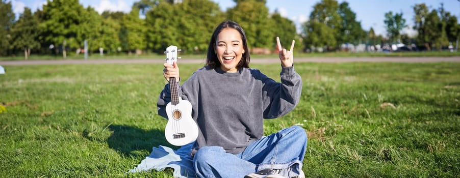 Positive korean girl sits in park, shows ukulele and rock on gesture, learns how to play musical instrument outdoors.