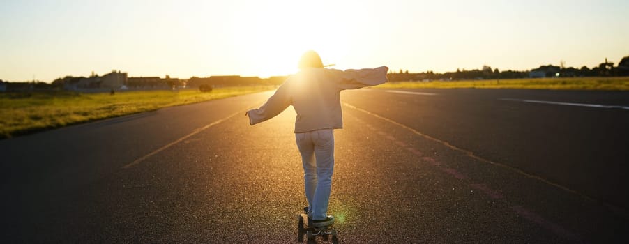 Rear view photo of young girl riding skateboard towards sunlight. Happy young woman on her cruiser, skating on longboard.
