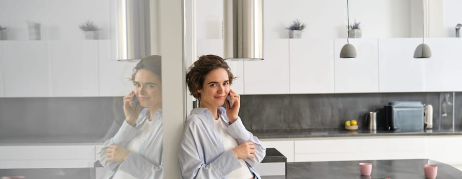 Beautiful woman talking on mobile phone, answers phone call and smiles, stands at home, leans to wall and chats.
