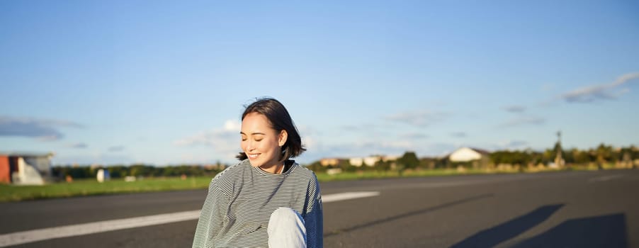 Cute smiling asian girl sits on longboard, skateboarding alone on an empty road. Copy space.