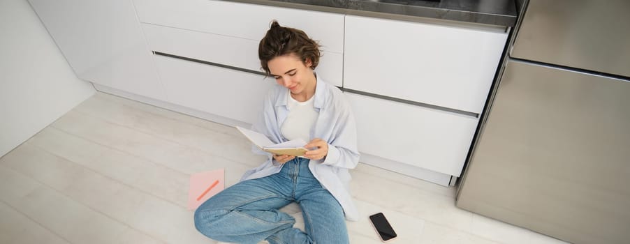 Upper angle shot of young woman sitting on kitchen floor, revising for exam, reading her notes, doing homework at home.