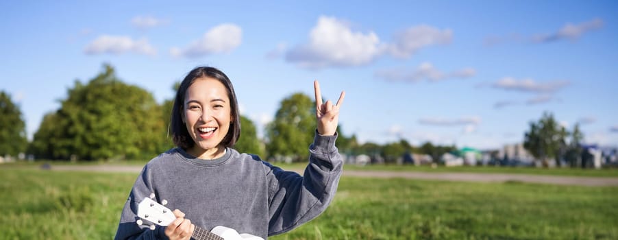 Happy asian girl playing ukulele in park, showing rock n roll, heavy metal horns sign and smiling, having fun outdoors.