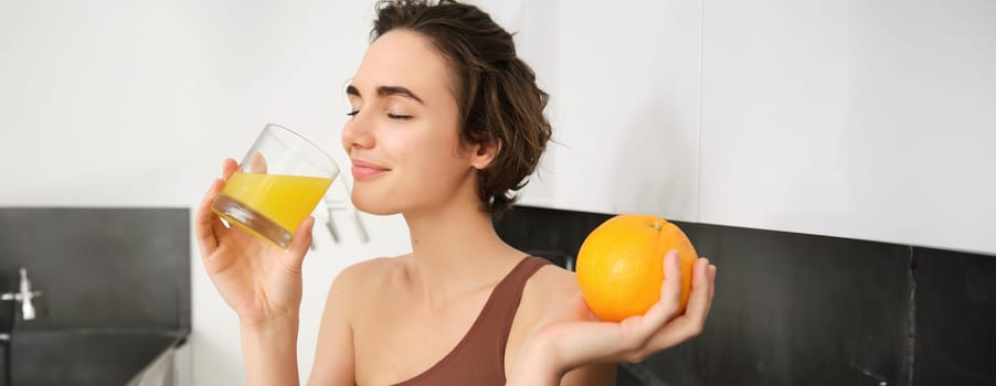 Healthy lifestyle and sport. Beautiful smiling woman, drinking fresh orange juice and holding fruit in her hand, enjoying her vitamin drink after workout at home, standing in kitchen.