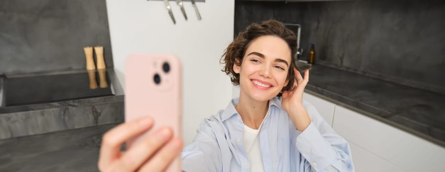 Happy, beautiful brunette girl takes selfie at home, poses for photo with smartphone.