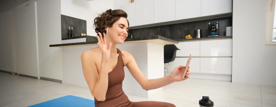 Young fitness girl at home, sport instructor in tracksuit waves hand at smartphone, sits on workout yoga mat, waves hand at mobile camera, says hi to online gym class, exercise remotely.