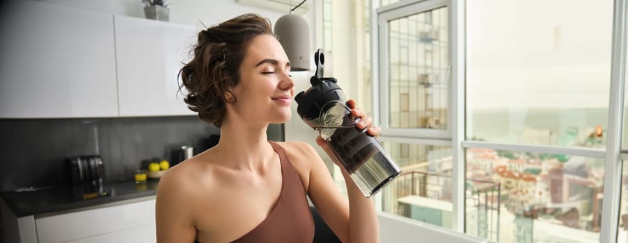 Sport and fitness concept. Portrait of young woman athlete drinking water from gym bottle, smiling with pleased face, wearing acrivewear, standing in kitchen.