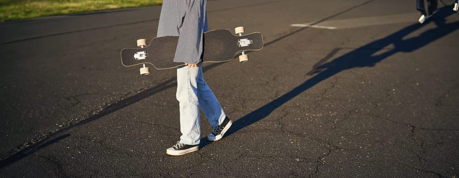 Cropped shot of teen girl body, holding cruiser longboard in hand, walking in sneakers on road in jeans and sweatshirt. Young woman skater with skateboard.