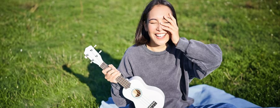 Smiling asian girl with ukulele, playing in park and singing, lifestyle concept. Copy space