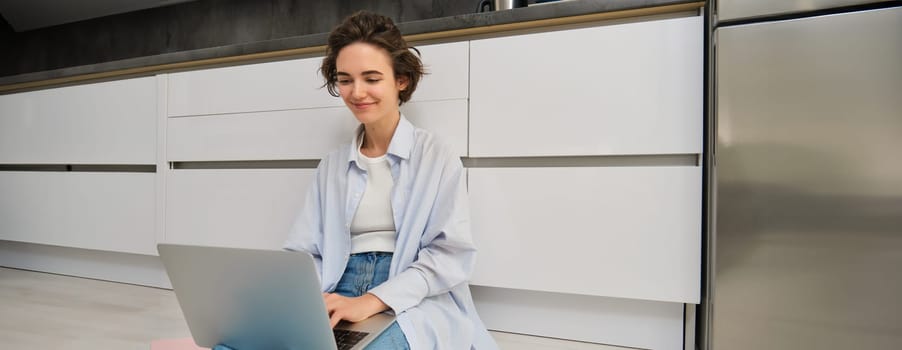 Young IT girl, woman works freelance from home, sits on floor with laptop. Girl student does homework online, writes on computer keyboard.