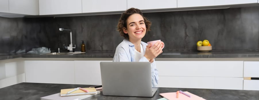 Smiling brunette girl sits in kitchen with laptop, drinks coffee and works from home.