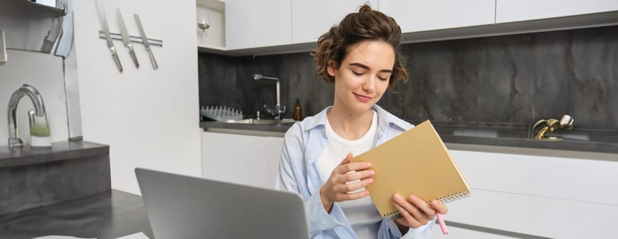 Young woman does homework, works from home, makes notes and uses laptop, managing business on remote, studies online.