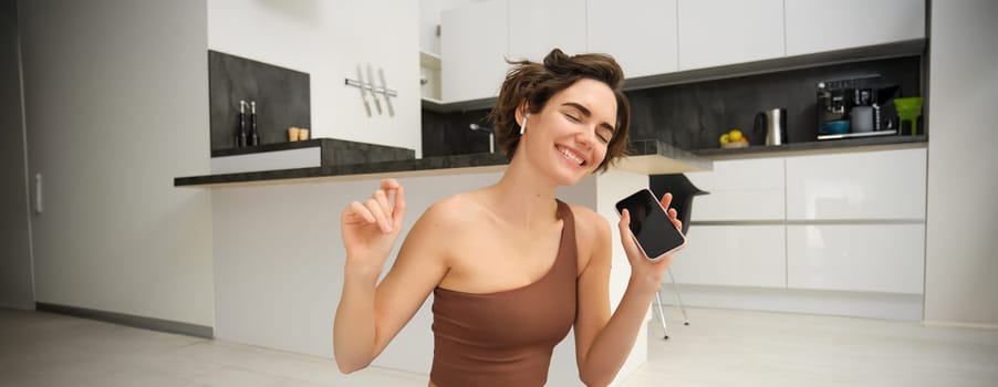Active lifestyle and health. Happy young woman does workout, sits and dances with smartphone in hands, listens music in wireless earphones during fitness training session, yoga on floor mat.