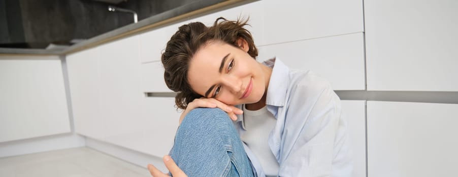 Portrait of brunette girl daydreaming, sitting on kitchen floor at home and smiling, looking aside with dreamy, thoughtful face expression.