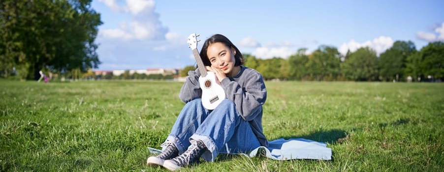 Cute romantic asian girl sitting with her instrument in park, hugging ukulele and smiling, resting outdoors.