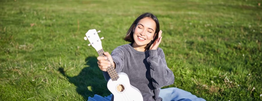 Smiling asian girl with ukulele, playing in park and singing, lifestyle concept. Copy space