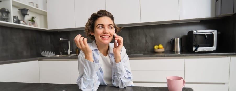 Cellular technology and people. Smiling brunette woman sits at home in kitchen, talks on mobile phone.