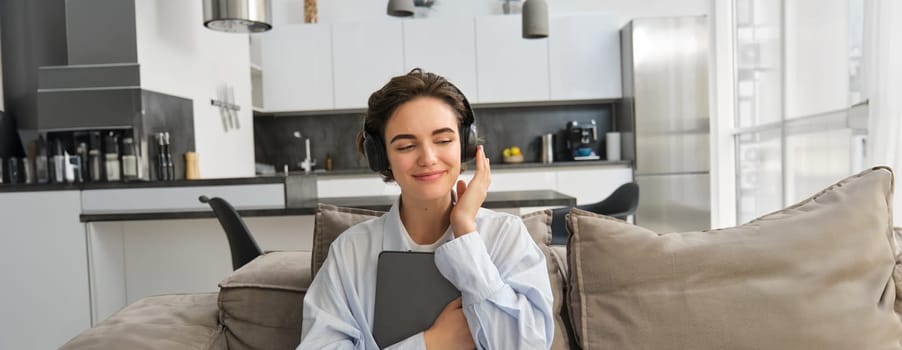 Portrait of brunette woman sits on sofa with tablet, listens music or podcast in headphones, enjoys ebook while spends time at home. Copy space