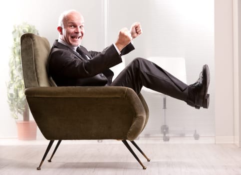 Cheering and exulting mature businessman on an armchair in an office laughing and looking at you