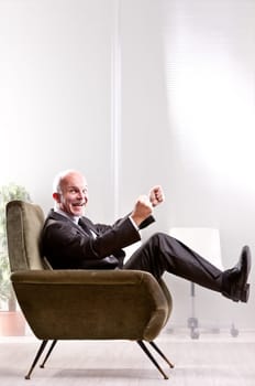 Cheering and exulting mature businessman on an armchair in an office laughing and looking at you