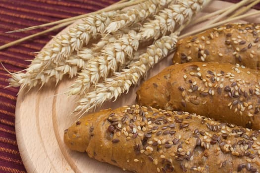 Wholemeal bread is  lying on a wooden tray on the table. Wheat ears are lying beside them.